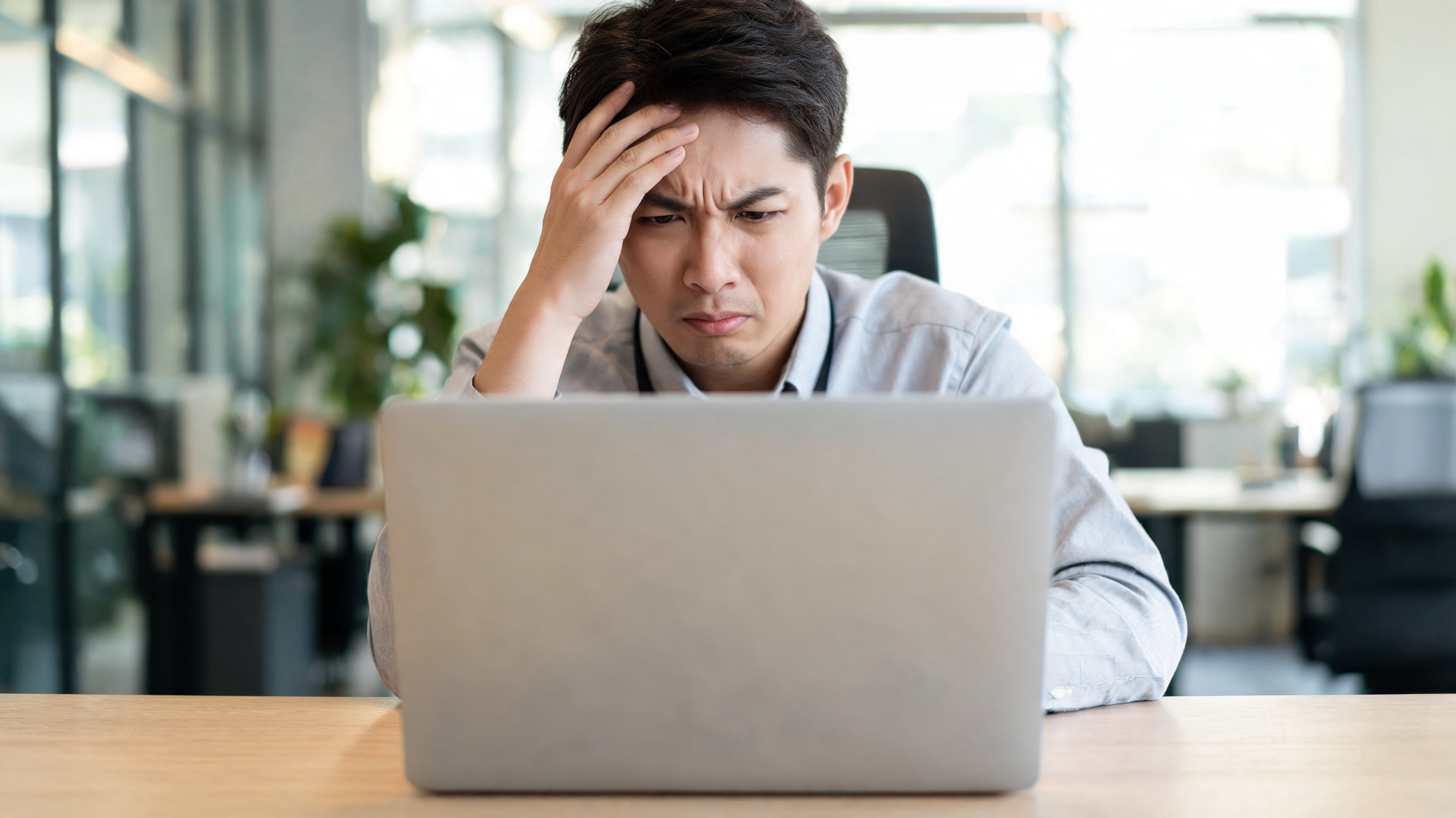 Frustrated customer experience manager at a desk in a modern office, hand on forehead, looking stressed behind a laptop.