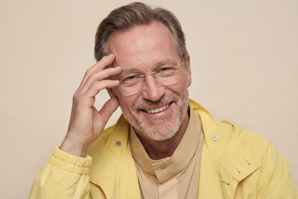 Smiling bearded man in a yellow jacket adjusting his rimless reading glasses with gold temples against a neutral studio background
