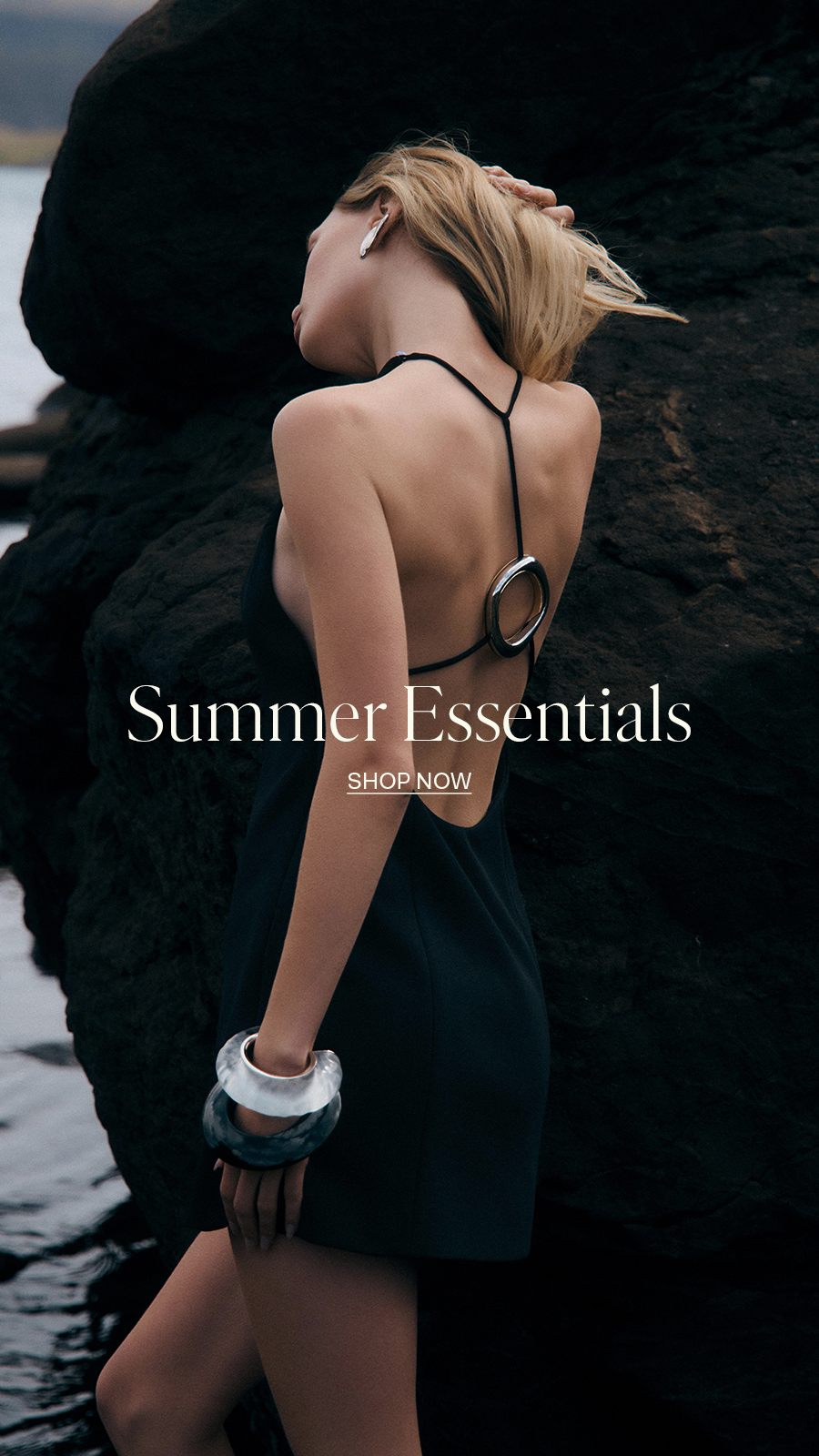 Woman in black backless halter dress with silver bangles posing near dark rocks, Summer Essentials fashion campaign