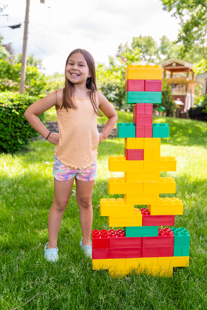 ```html
<img src="image.png" alt="Child standing next to a tall tower made of large colorful plastic blocks in a grassy backyard." />
```
