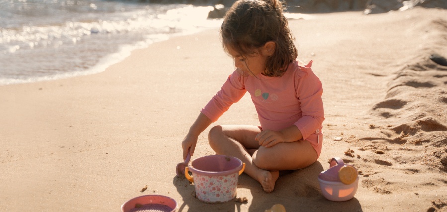 Young girl in a pink swimsuit sitting cross-legged on a sandy beach, playing with toy buckets near the water's edge.