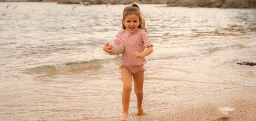 Young girl in pink swimsuit running along sandy beach shoreline, holding a small ball, smiling