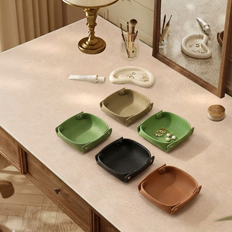 Six small leather catchall trays in tan, green, black, and brown colors arranged on a vanity dresser with a mirror and makeup brushes.