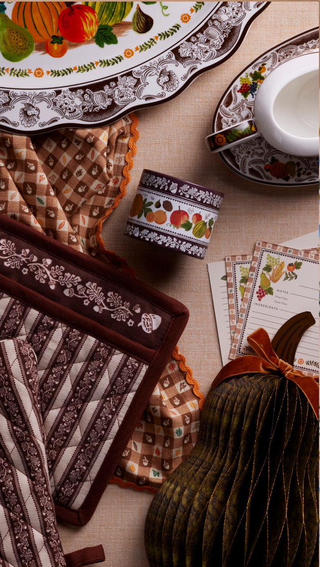 Autumn-themed kitchen items including decorative plates, pot holders, recipe cards, washi tape, and a velvet pumpkin decoration arranged on linen fabric.