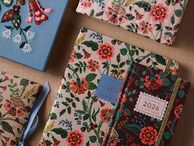 ```html
<img src="image.jpg" alt="Collection of floral patterned planners and notebooks, some labeled with 2026, arranged on a brown surface." />
```