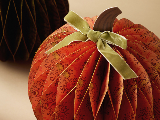 Decorative paper pumpkin with fall harvest print, green velvet bow, and wooden stem, with dark honeycomb decoration in background.