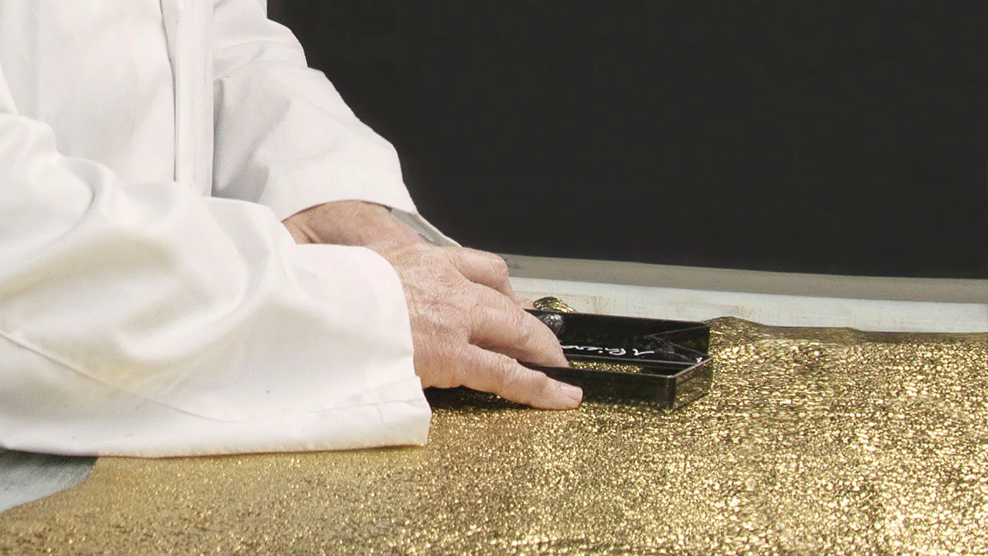 Close-up of a person in a white lab coat holding a black rectangular object over a surface covered in gold glitter.