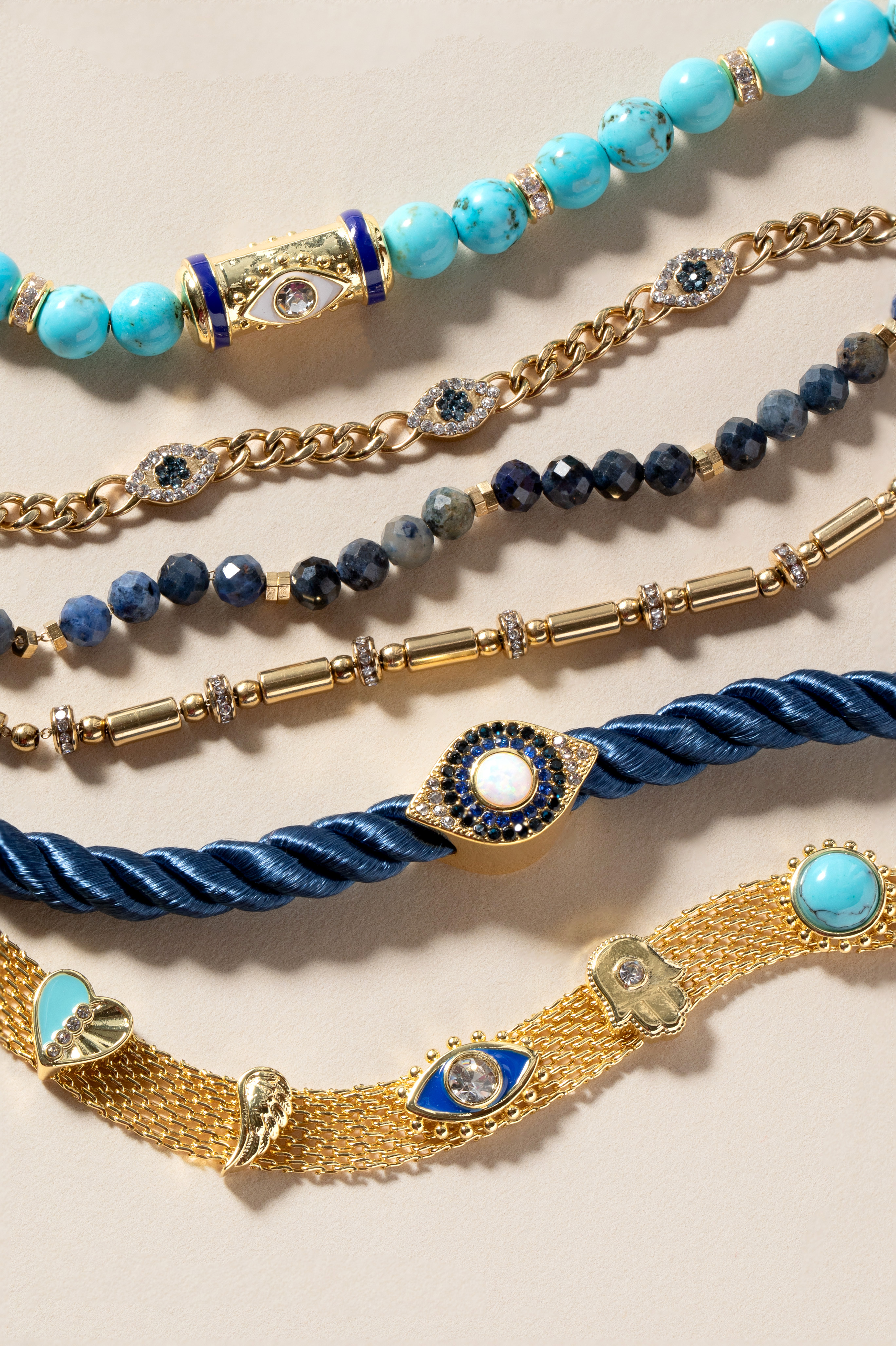 Five layered bracelets featuring evil eye charms in gold, turquoise beads, blue gemstones, twisted rope, and mesh chain designs.