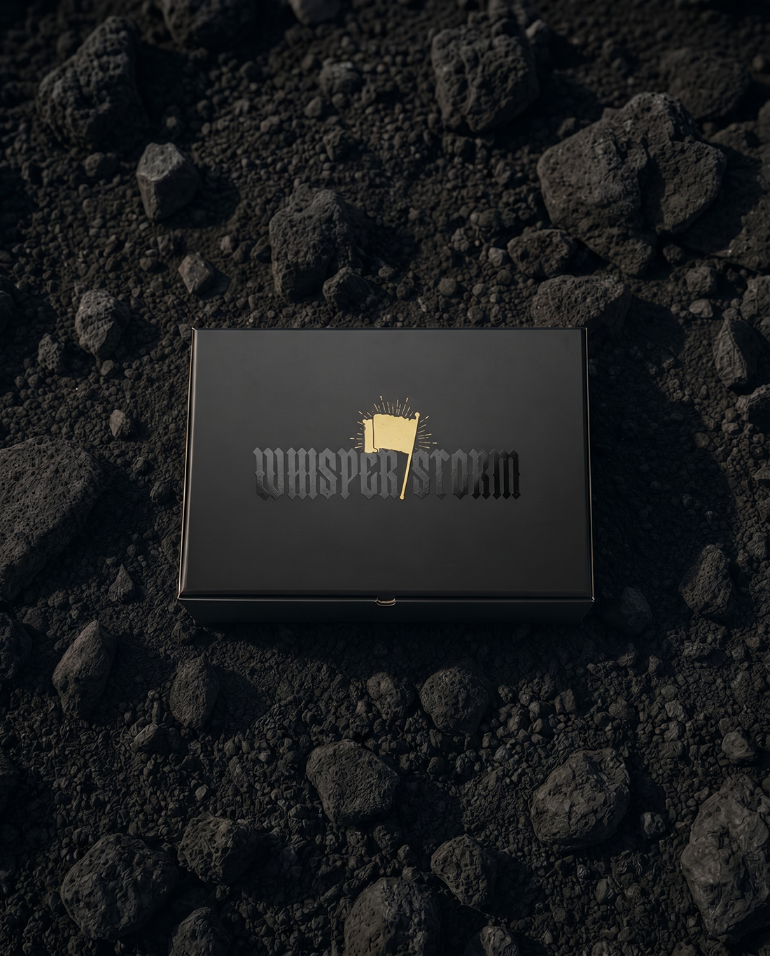 Black rectangular gift box with gold "Whisper Storm" logo resting on dark volcanic rocks and gravel