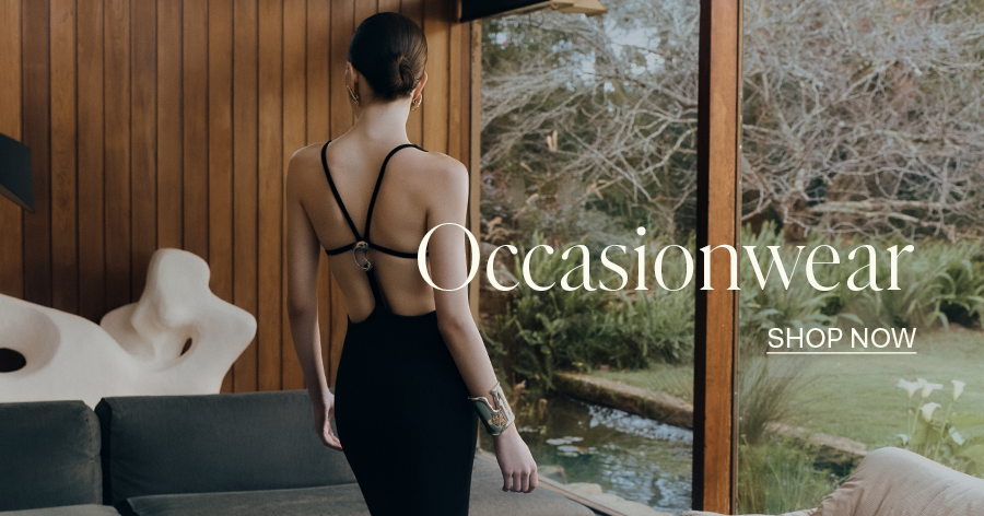 Woman in black open-back occasionwear dress standing near floor-to-ceiling windows with garden view, "Occasionwear - Shop Now" text overlay