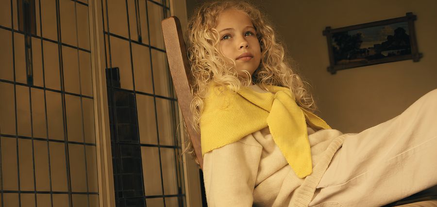 Young girl with curly blonde hair sitting in a wooden chair, wearing a yellow top, looking upward in a warmly lit room.