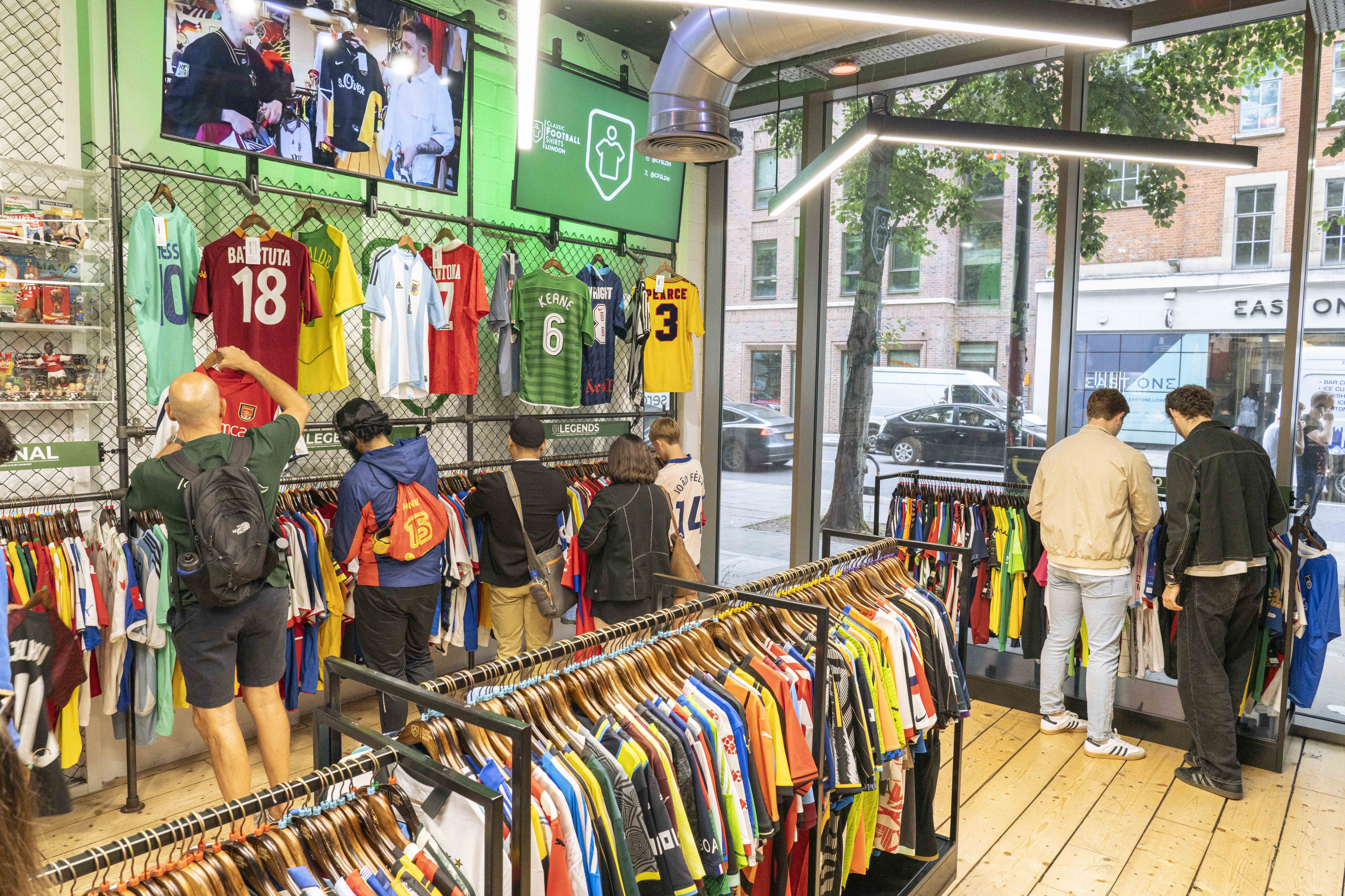 ```html
<img src="image.jpg" alt="People browsing football jerseys in a sportswear store, with various colorful jerseys displayed on racks and walls. A screen shows a video, and a street view is visible through large windows." />
```