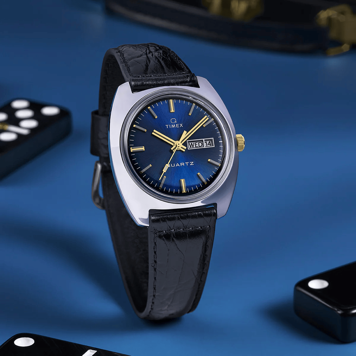 ```html
<img src="image.jpg" alt="Timex wristwatch with blue dial, black leather strap, and gold accents, displaying day and date, on a blue surface with dominoes nearby">
```