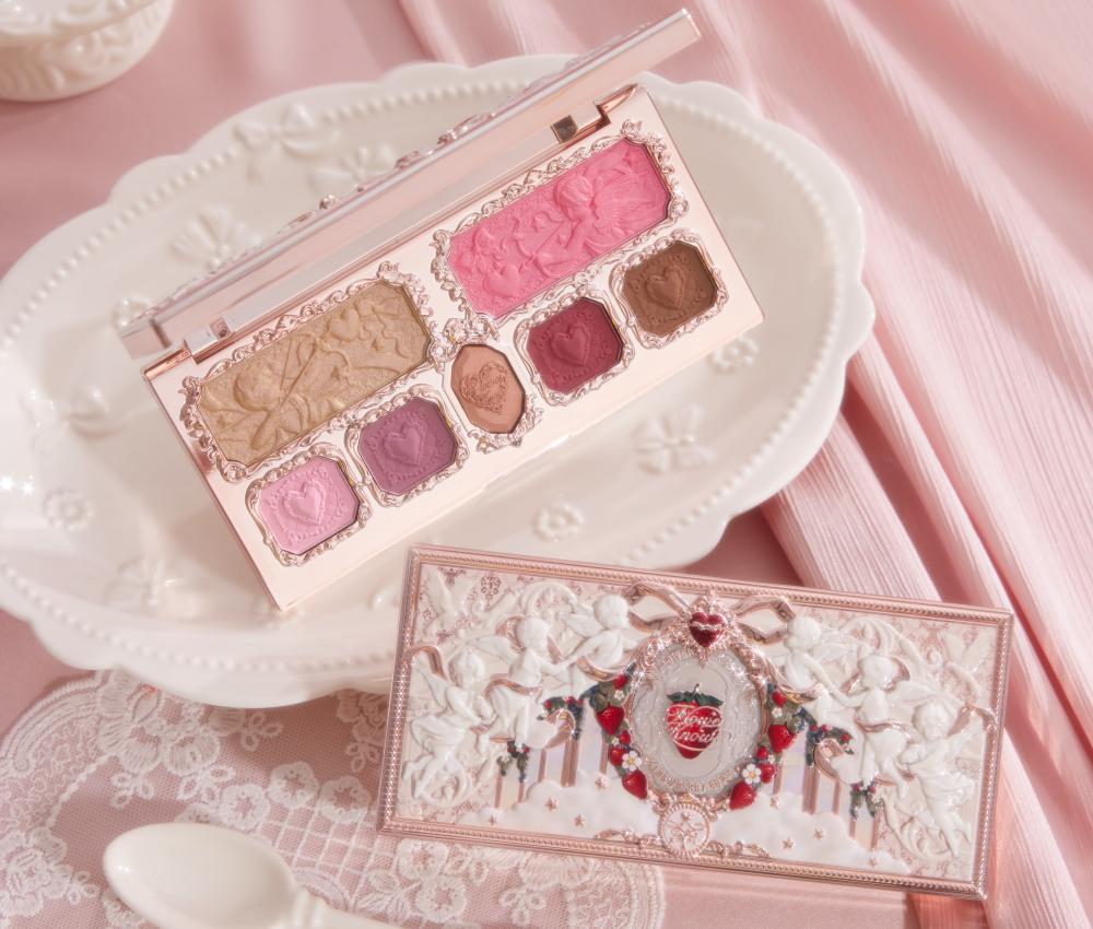 <alt text>Makeup palette with decorative packaging on a white plate and pink fabric background. The palette includes various shades of eyeshadow.</alt text>