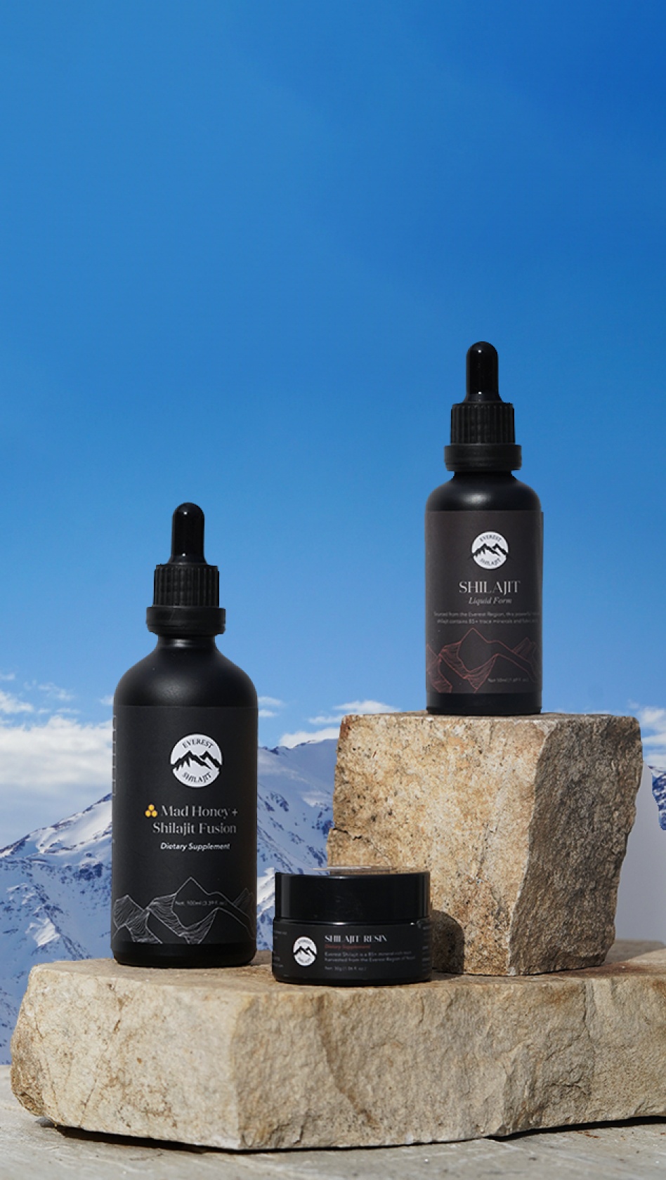 Three black bottles of Shilajit products are displayed on rocky surfaces with a clear blue sky and snow-capped mountains in the background.