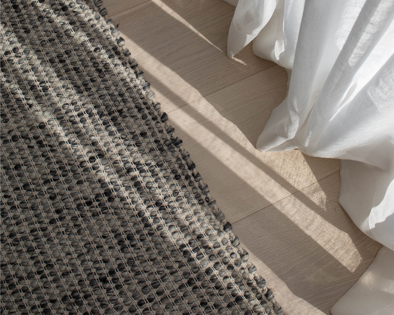 Close-up of a gray woven textured rug on light wood floor beside a white sheer curtain with sunlight shadows