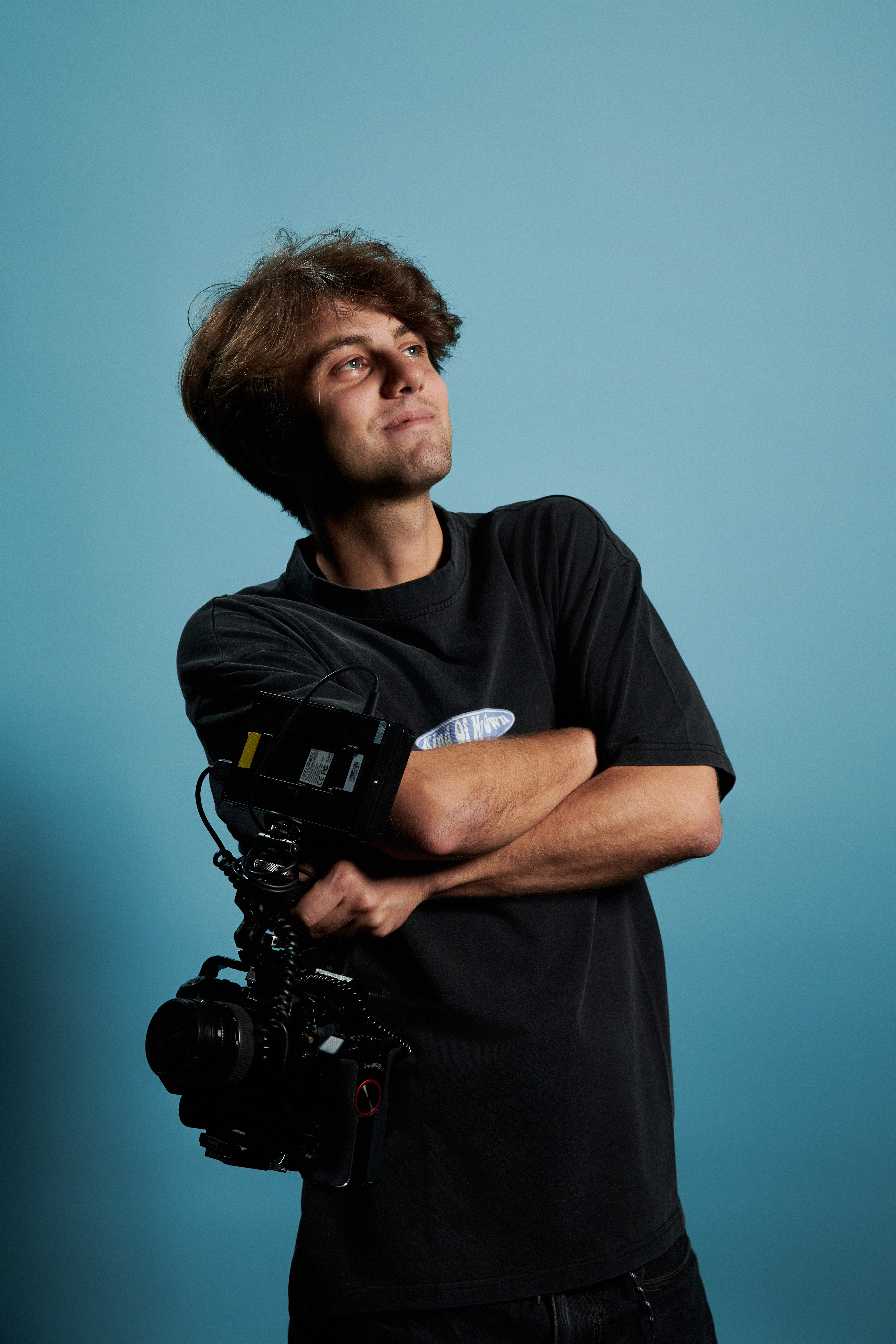 Person in a black t-shirt holding a camera with a large lens and attached equipment, standing against a blue background.