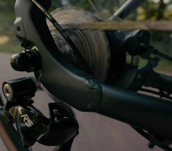 Close-up of a bicycle rear derailleur and spinning wheel hub on a dark-framed bike in motion outdoors.