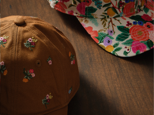 Two baseball caps on a wooden surface; one brown with small floral embroidery, the other white with a colorful floral print.