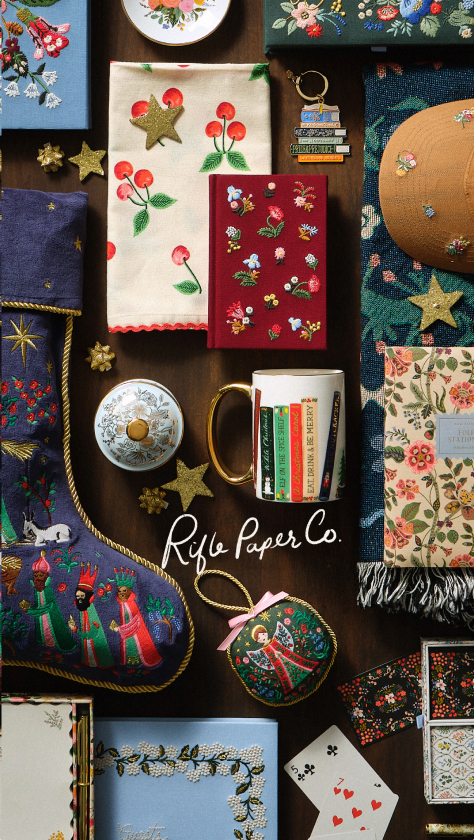 A collection of Rifle Paper Co. products on a wooden surface, including floral notebooks, a mug, decorative plates, a keychain, cards, and a fabric tote.