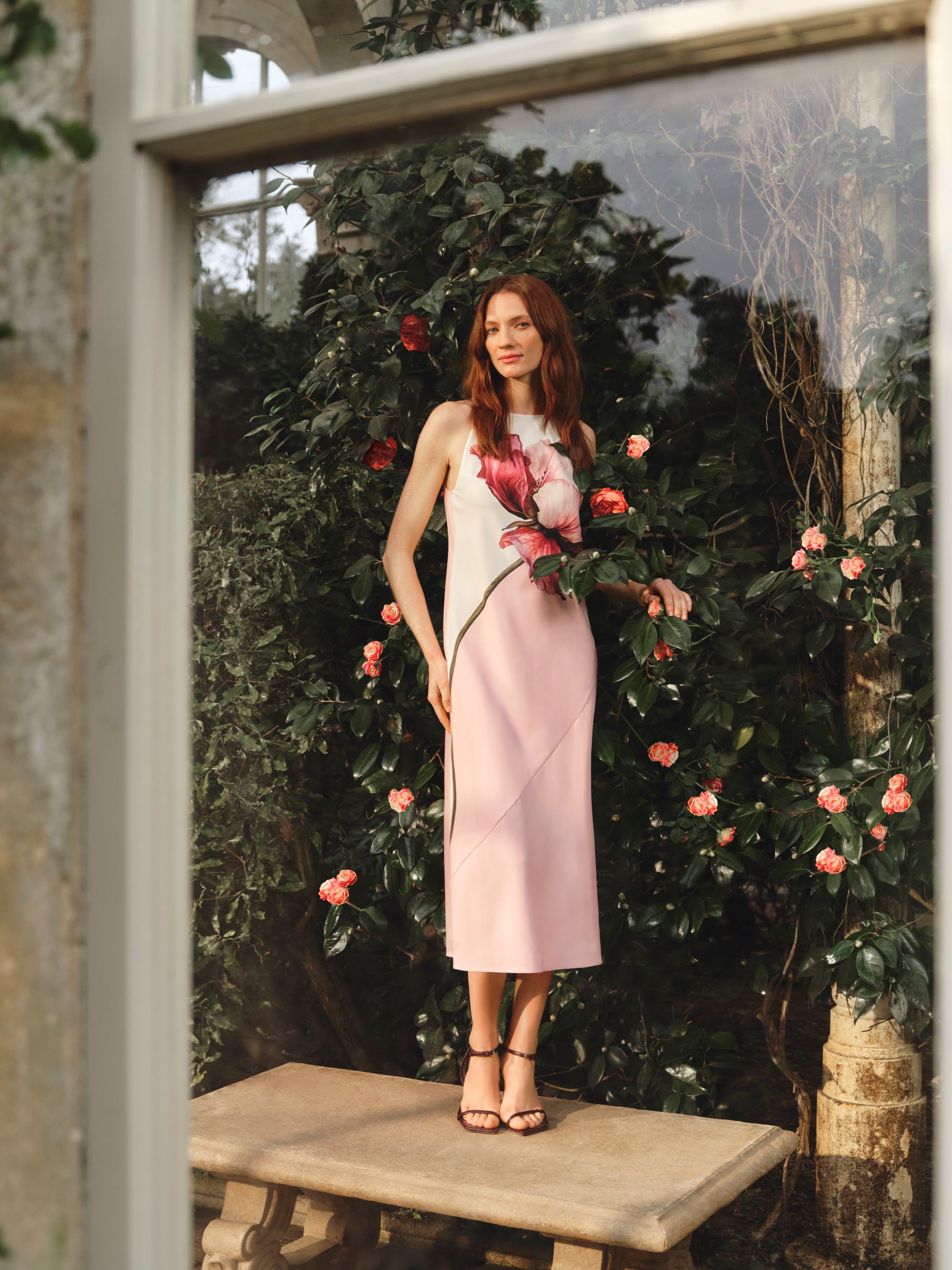 ```html
<img src="image.jpg" alt="Person in a pink dress standing on a stone bench surrounded by flowering bushes, viewed through a window." />
```