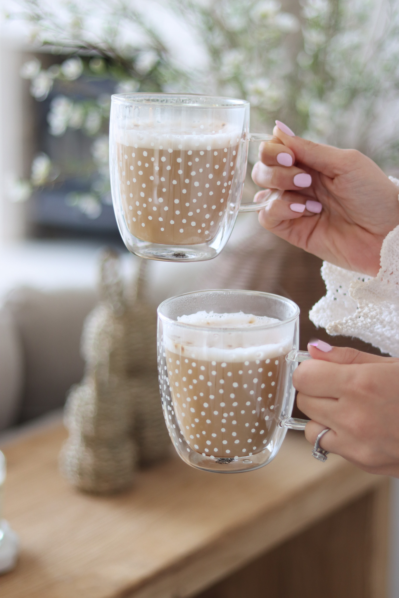 Two hands holding glass mugs with polka dot designs, containing frothy coffee. Blurred background with greenery.