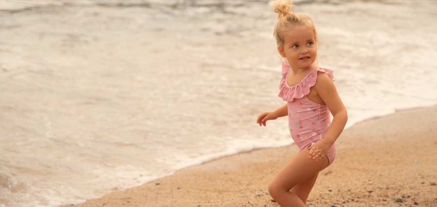 Young toddler girl in pink ruffled swimsuit running on sandy beach with ocean waves in background