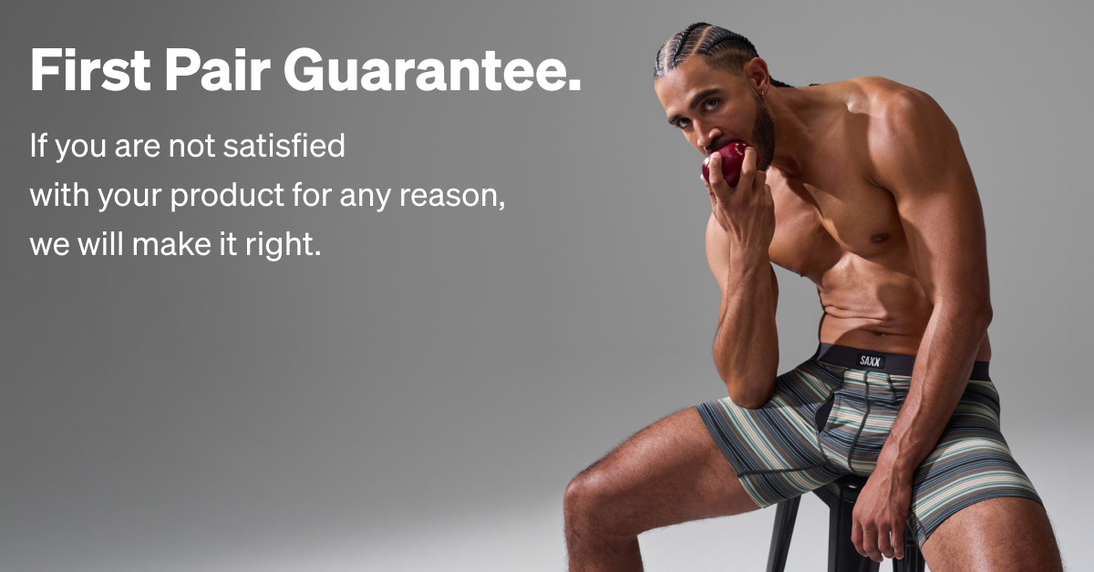Male model sitting on stool wearing SAXX striped boxer briefs, holding apple, with "First Pair Guarantee" promotional text on gray background.