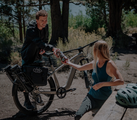 Two cyclists resting outdoors, man handing a drink bottle to a seated woman beside a cargo e-bike, helmets nearby.