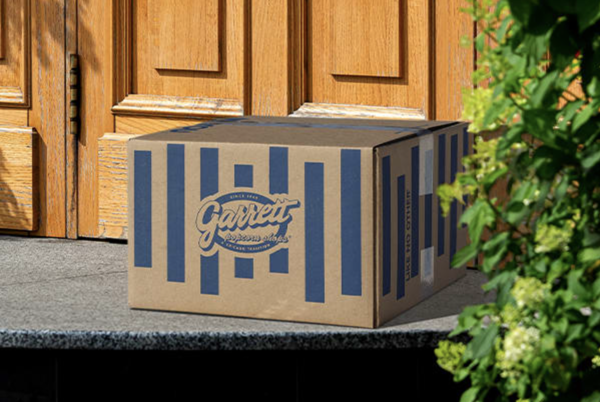 Garrett Popcorn Shops branded cardboard delivery box with blue stripes, placed on a front doorstep near a wooden door.