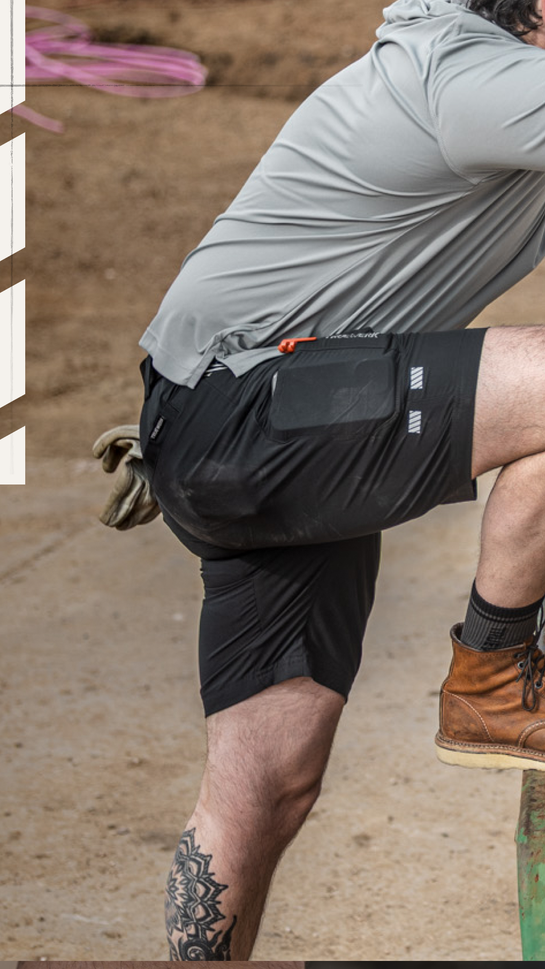 Man wearing gray long-sleeve shirt, black work shorts with utility pockets, brown leather boots, and work gloves, with leg tattoo visible