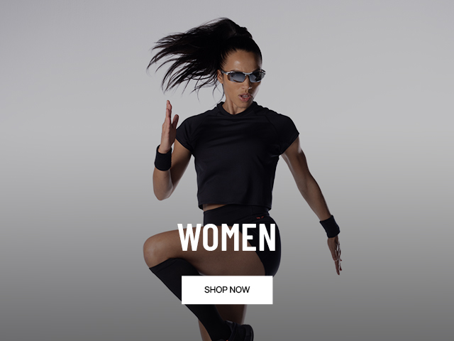 A woman in athletic attire, including a black t-shirt, shorts, and knee-high socks, performs a dynamic jump. The image has "WOMEN" and "SHOP NOW" text overlay.