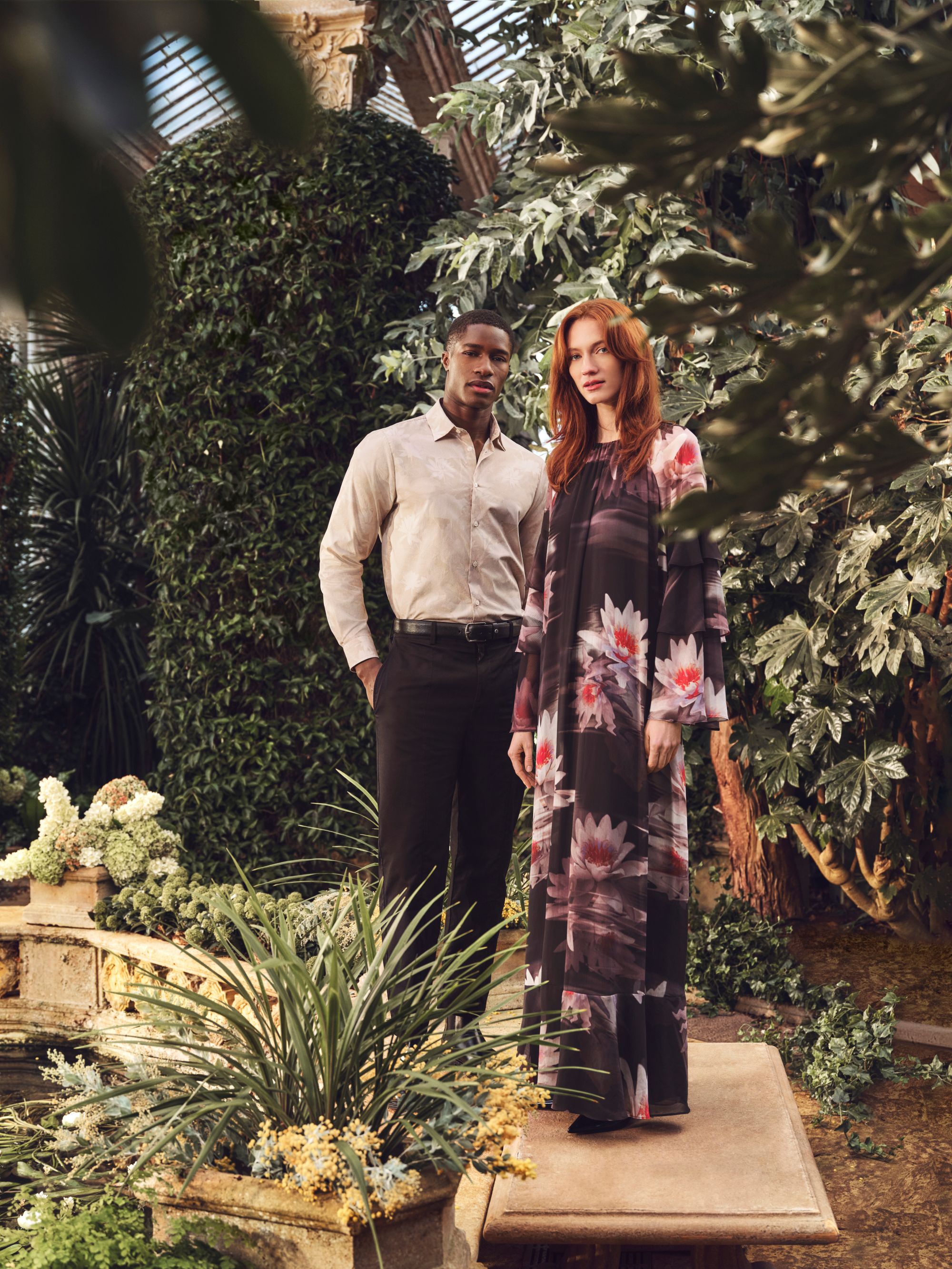 A man in a light shirt and dark pants stands next to a woman wearing a floral dress. They are in a lush indoor garden with various plants and greenery.