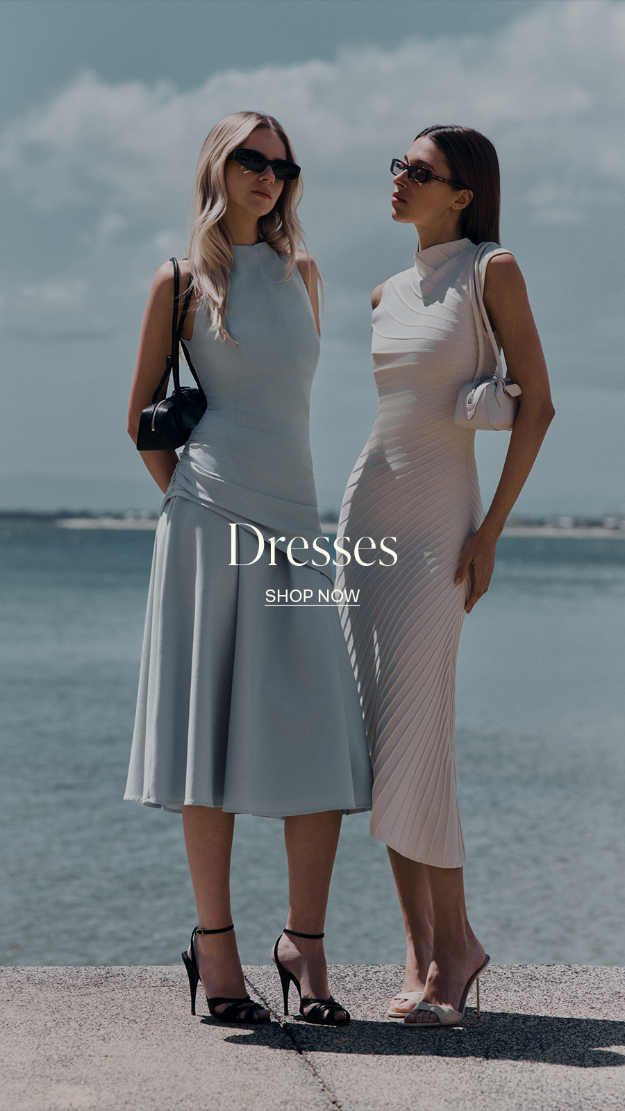 Alt text: Two women wearing elegant dresses stand outdoors by the water. One wears a light blue dress and carries a black bag, while the other wears a light pink dress and holds a matching clutch. Text overlay reads "Dresses - Shop Now."