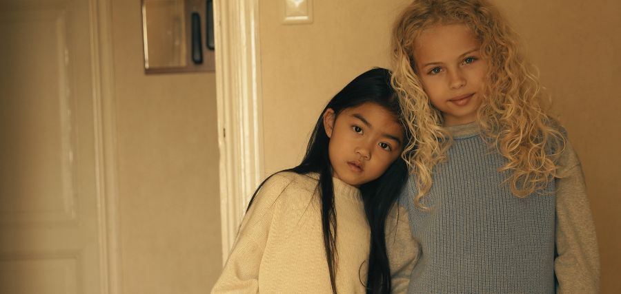 Two young girls posing together indoors, one with long dark hair in a cream sweater, the other with curly blonde hair in a blue top.