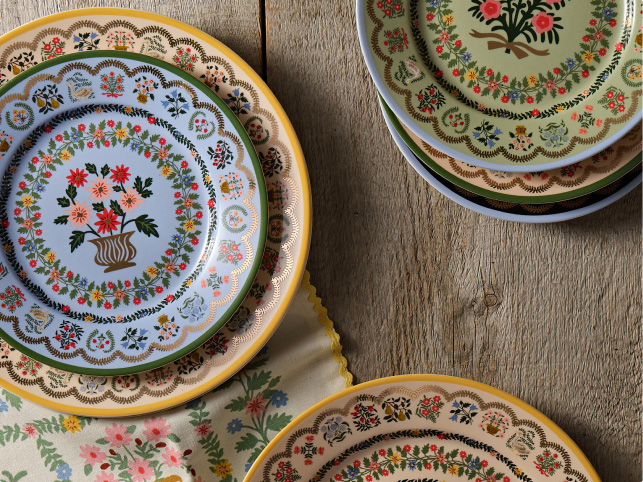 Decorative ceramic plates with colorful floral patterns and scalloped gold edges arranged on a rustic wooden surface with a floral cloth.