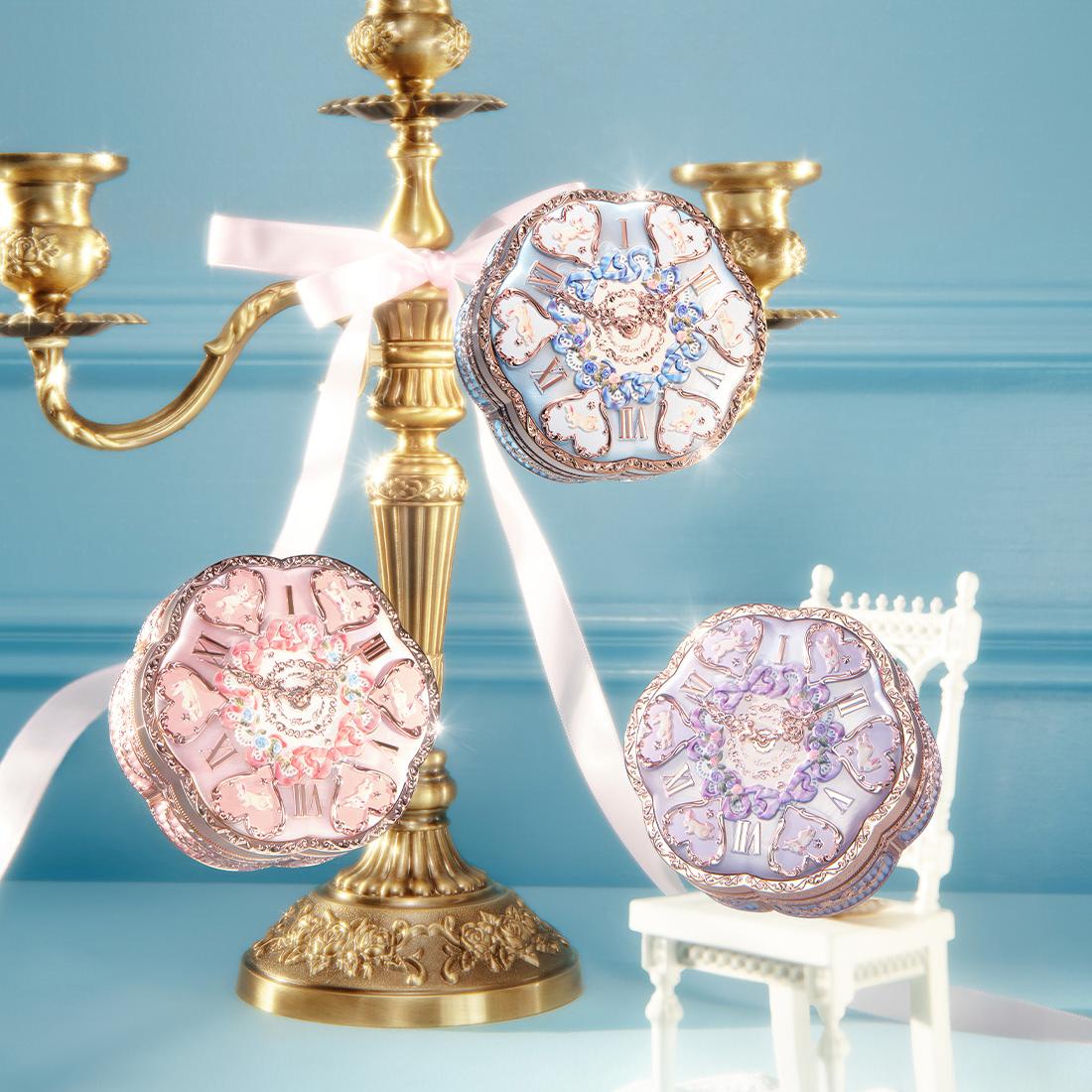 Three ornate clock-face compact mirrors in pink, blue, and purple with floral designs, displayed near a gold candelabra with white ribbons.