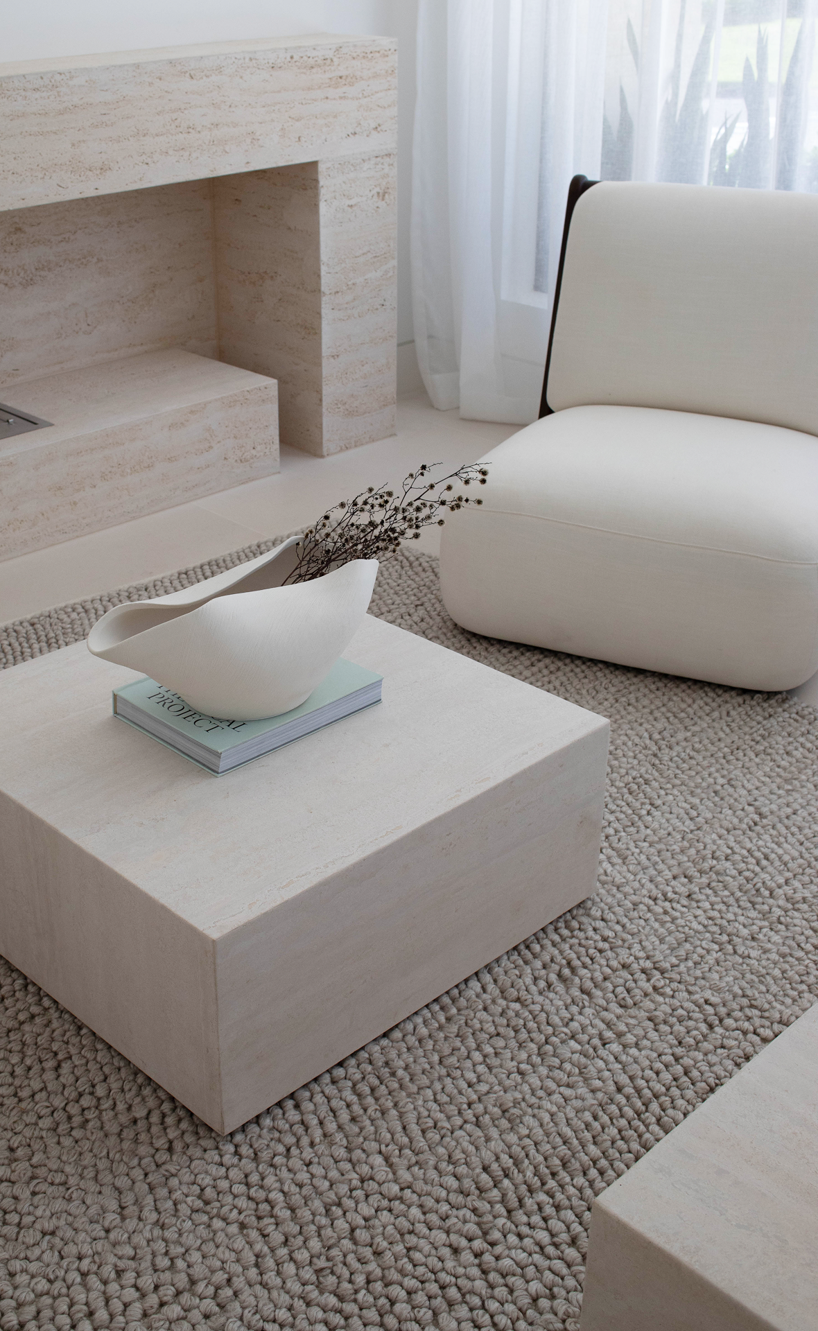 Minimalist living room with travertine stone coffee table, white sculptural bowl, dried branches, and cream upholstered chair on textured rug.