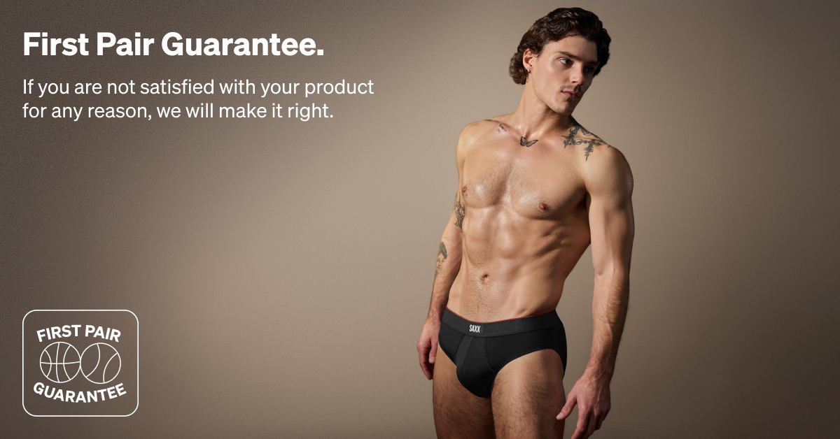 Alt text: A muscular man with tattoos wearing SAXX underwear stands against a neutral background. Text on the left reads "First Pair Guarantee. If you are not satisfied with your product for any reason, we will make it right."