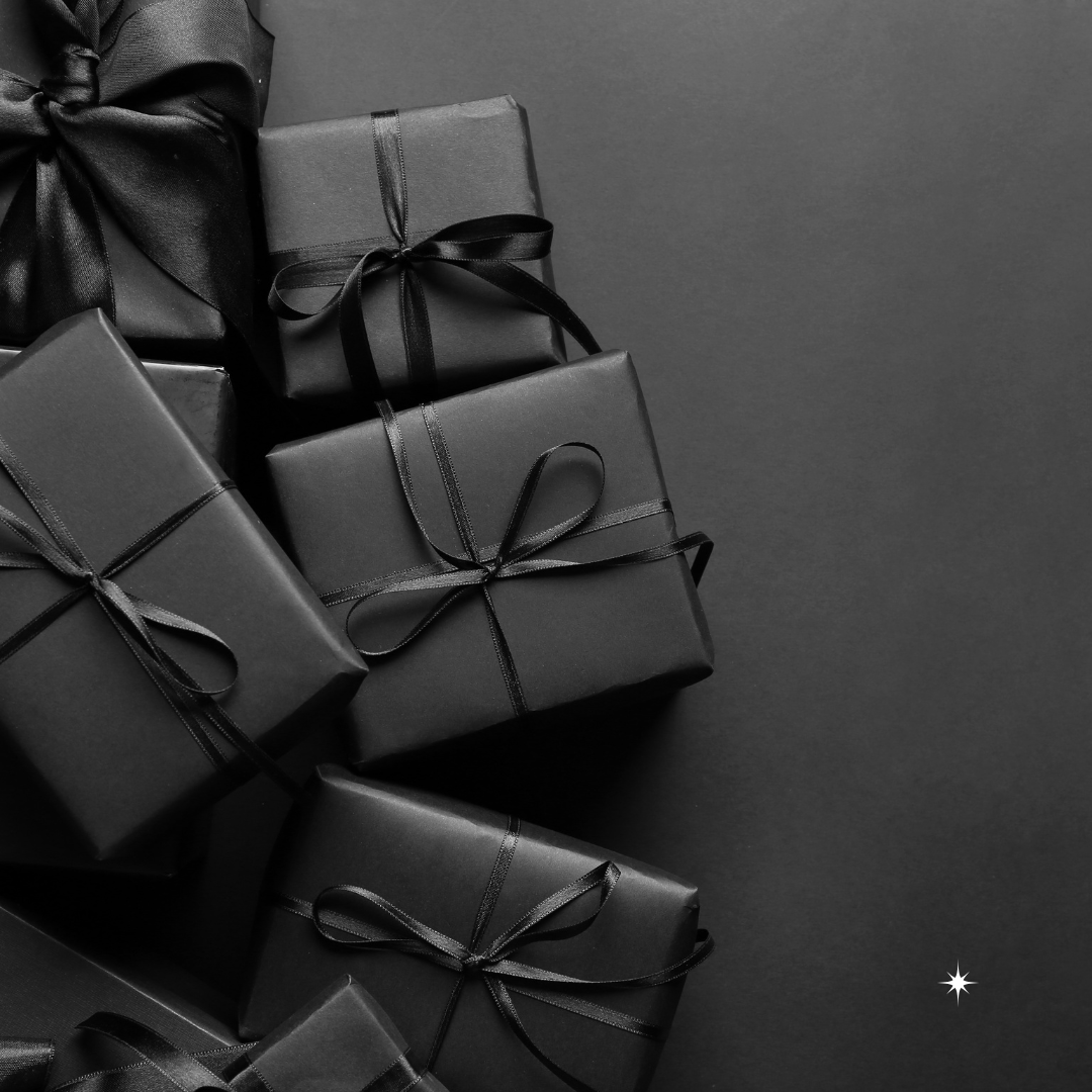 Several black gift boxes with black ribbons are stacked on a dark surface, with a small white star in the bottom right corner.