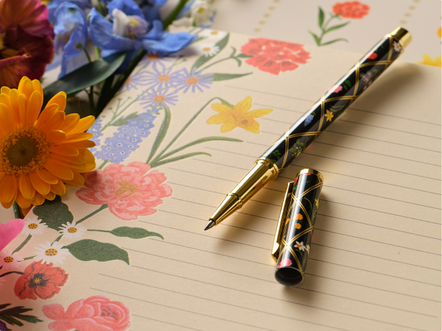 A floral-patterned fountain pen with gold accents rests on a lined notepad with colorful flower illustrations, surrounded by fresh blooms.