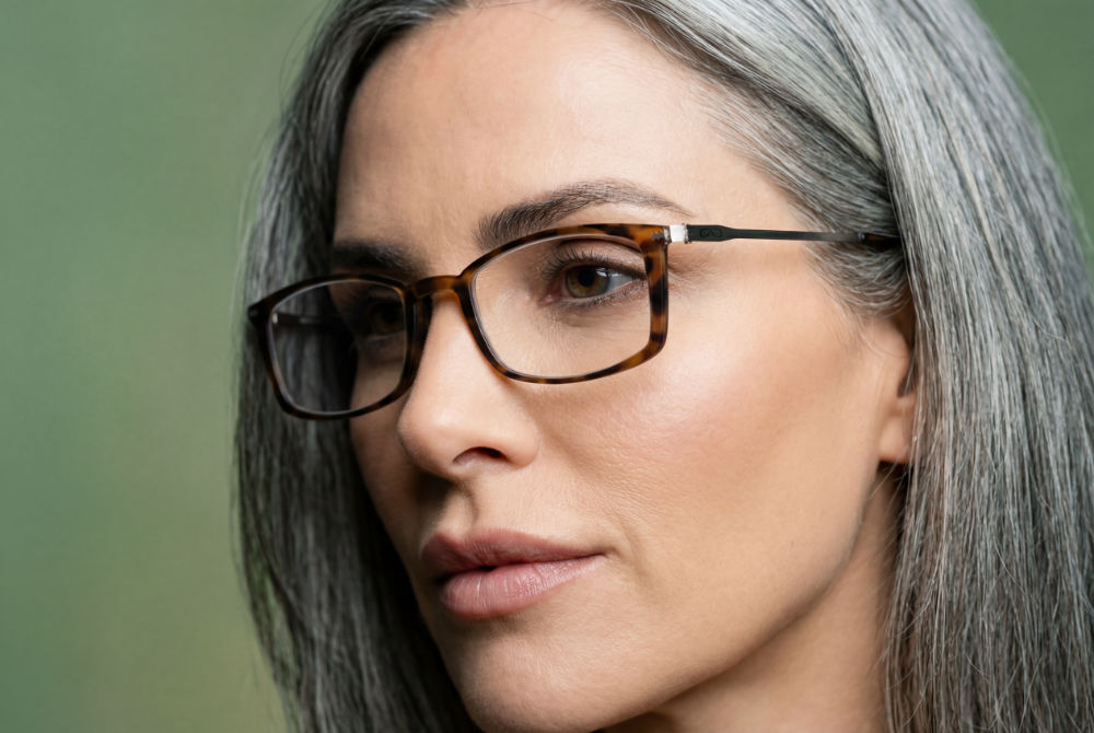 Close-up portrait of a silver-haired woman wearing tortoiseshell rectangular reading glasses with slim metal temples against a soft green background