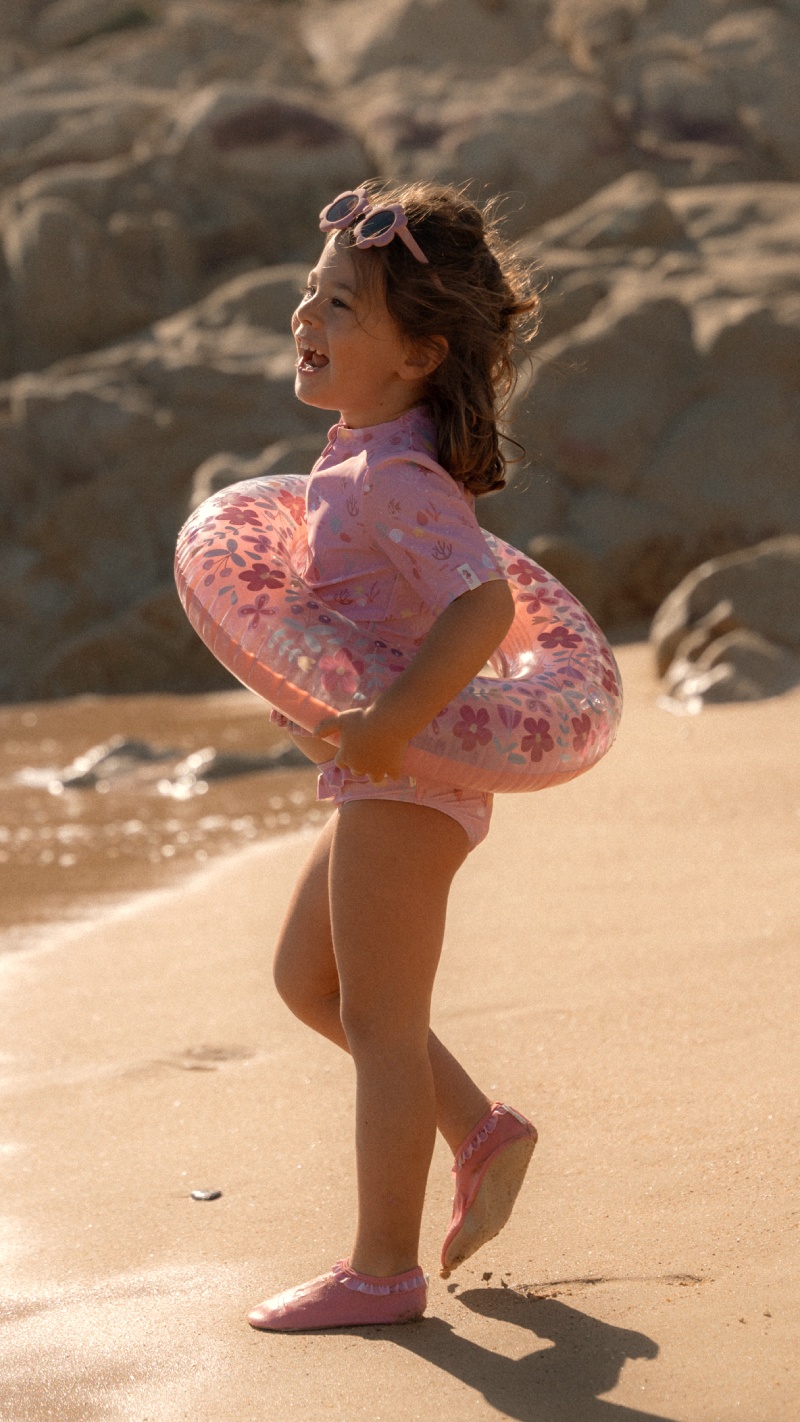 Young girl in pink swimsuit holding a floral inflatable ring on a sandy beach, wearing sunglasses on her head.