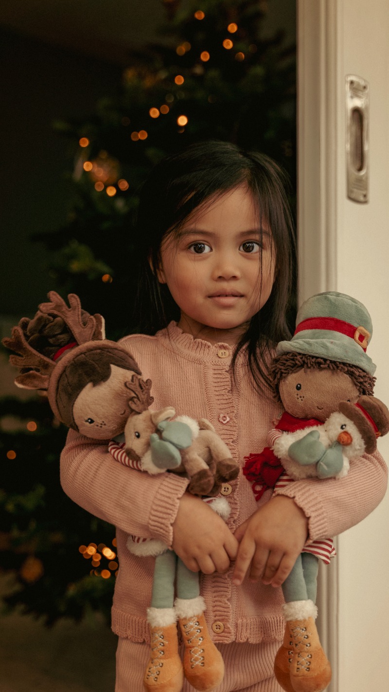 Child in a pink sweater holding festive plush toys, including a reindeer and snowman, standing near a Christmas tree with blurred lights in the background.