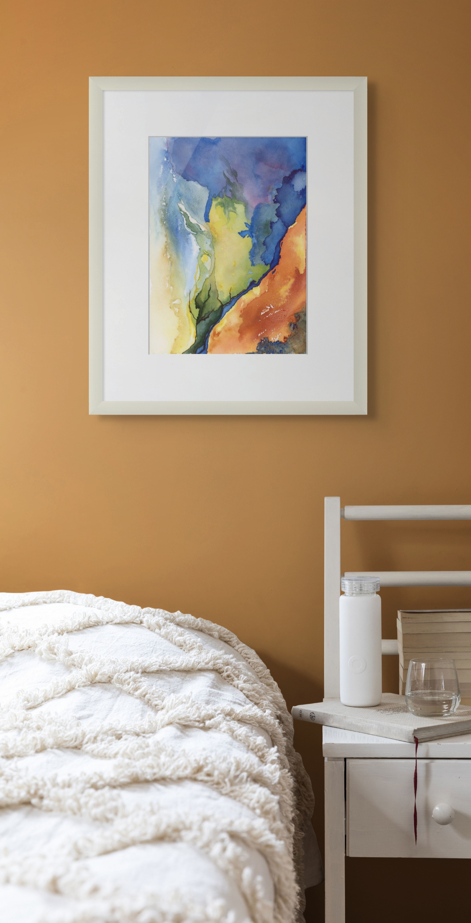 Framed abstract watercolor painting on a brown wall above a bed with white bedding. A white bedside table holds books, a glass, and a white bottle.