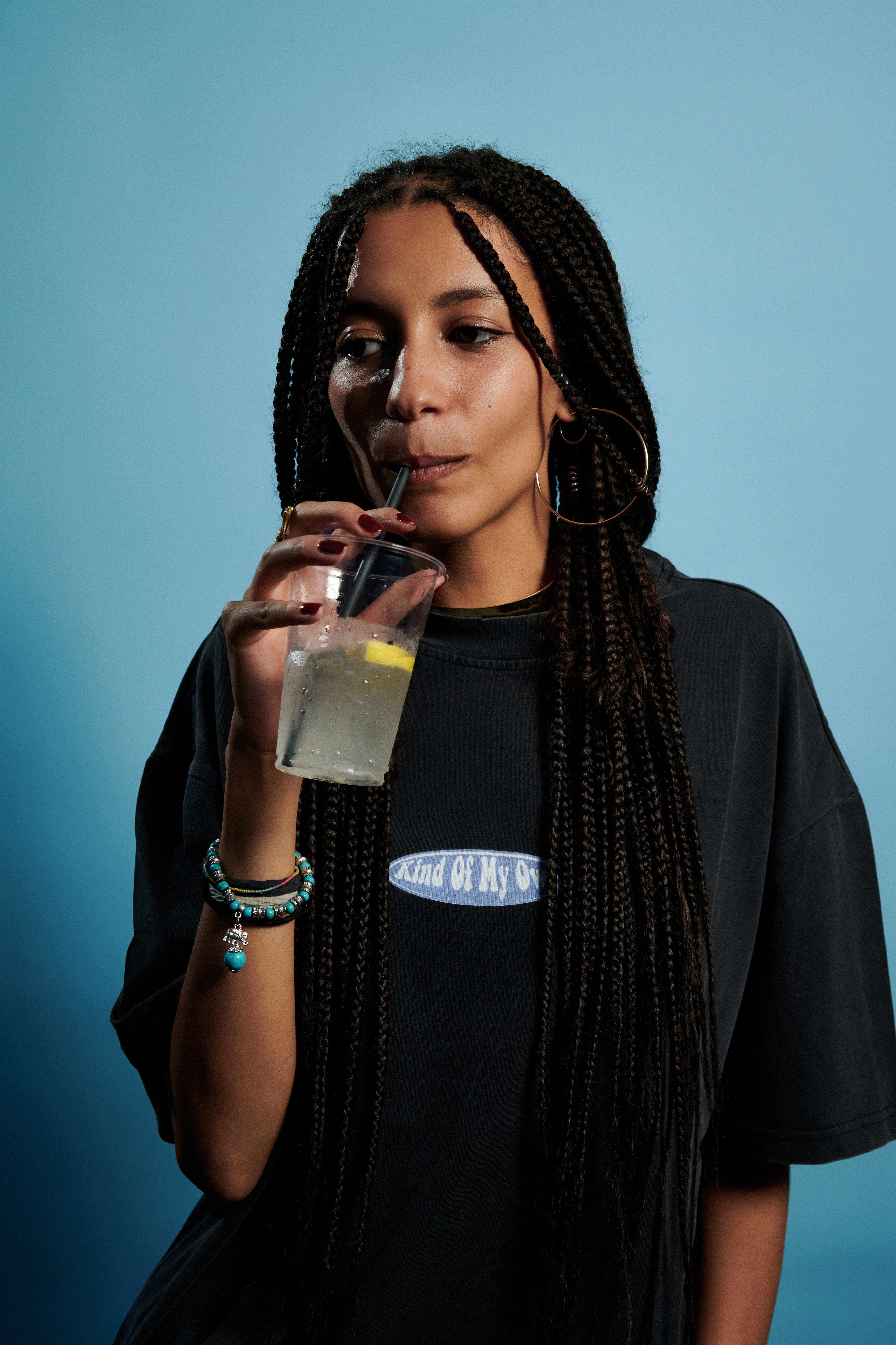 A person with long braided hair wearing a dark t-shirt and bracelets holds a plastic cup with a lemon slice against a blue background.