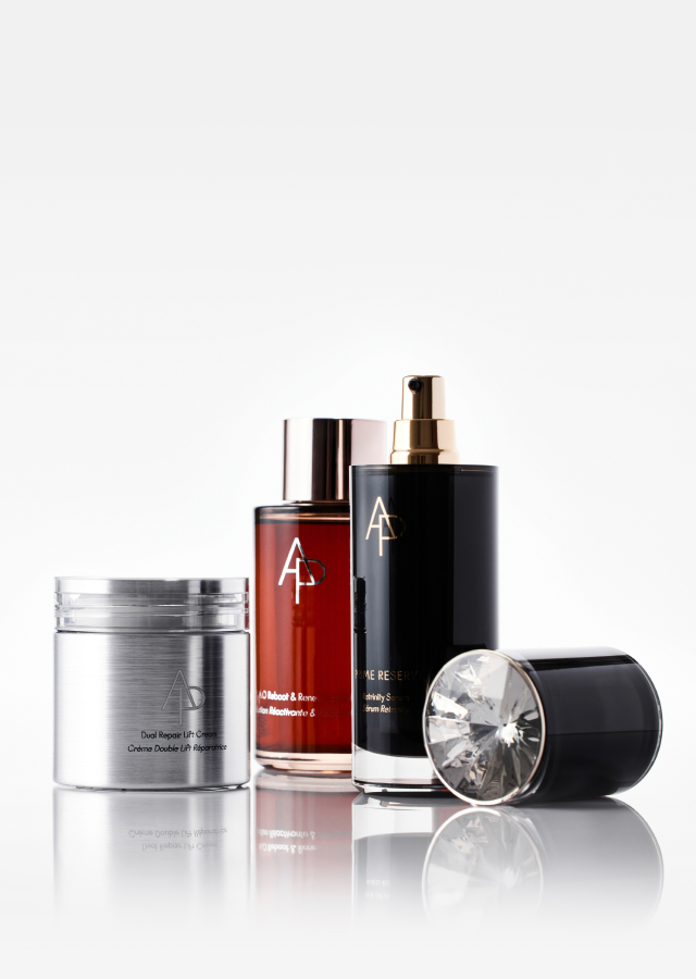 Three luxury skincare products with "AD" branding: a silver cream jar, a red bottle with rose gold cap, and a black serum bottle with gold pump.