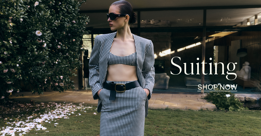 Woman in houndstooth suit set with crop top, blazer, and skirt, wearing sunglasses and black belt, "Suiting-  Shop Now" text overlay.