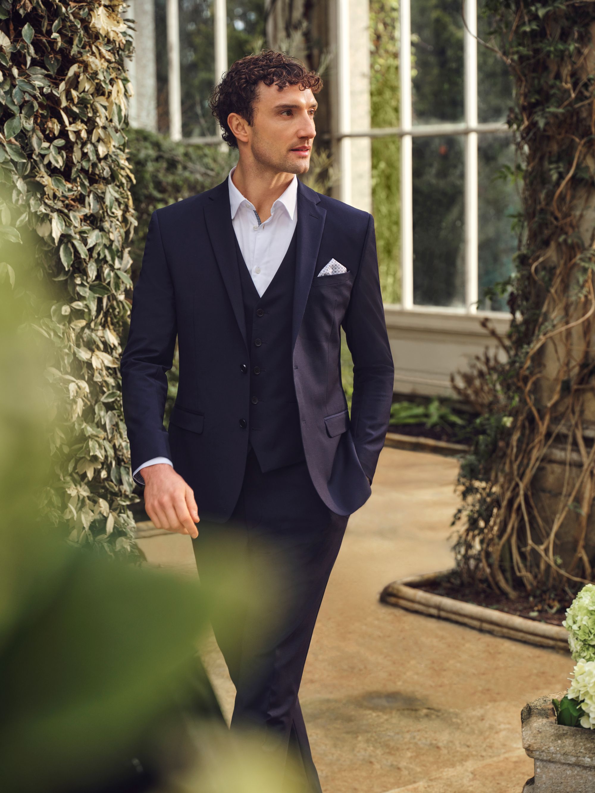 A person in a dark suit with a white shirt and pocket square stands outdoors, surrounded by greenery and a greenhouse structure.