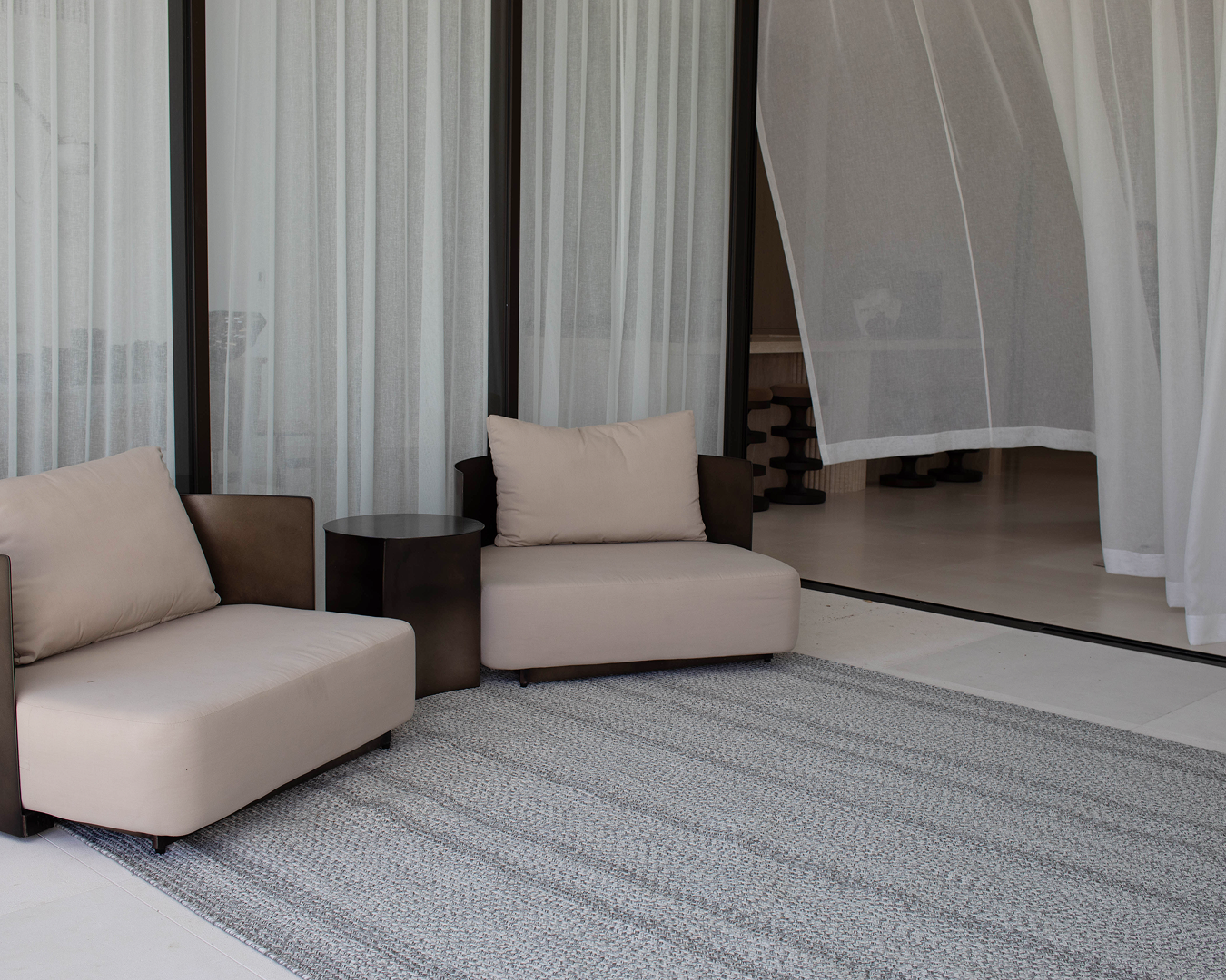 Two beige armchairs with cushions and a dark side table on a gray rug, framed by sheer white curtains.